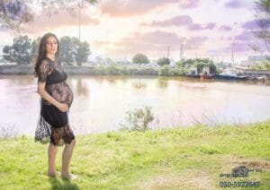 תמונה מתוך צילומי הריון. אישה הריונית עם שמלה שחורה עומדת על כר דשא על שפת הנחל ואוחזת בבטן ההריונית