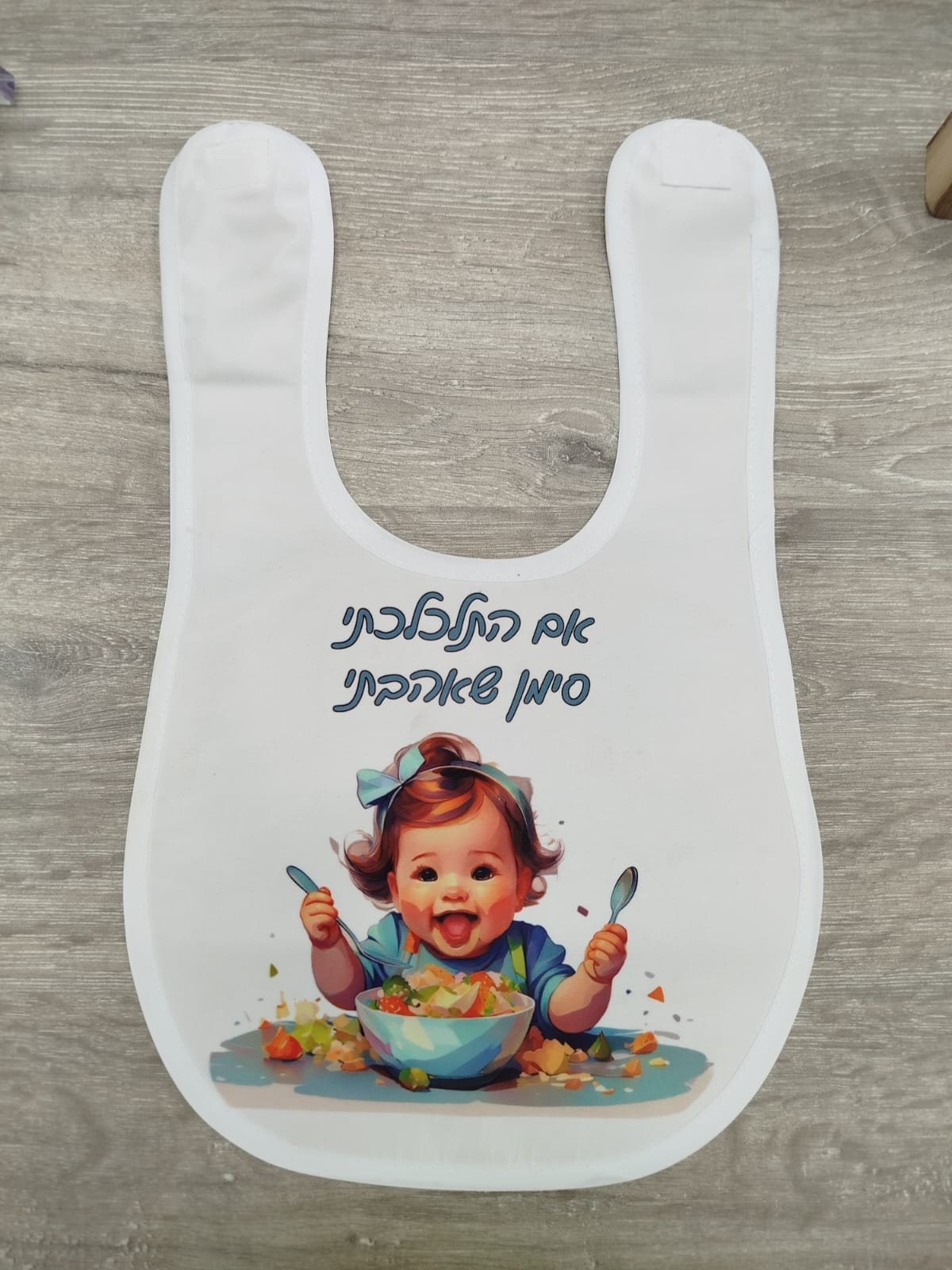 סינר לתינוק סינר לבן לתינוק עם הדפסה אישית הכוללת איור של תינוקת אוכלת והכיתוב בעברית "אם התלכלכתי סימן שאהבתי", מונח על שולחן עץ – מתנות בעיצוב אישי בהראל צלמים בקריות